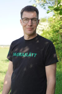 Per Modig, växtodlingsstrateg, Ekologiska Lantbrukarna. Halvfigur i halvskugga av en buske. på en svart t-shirt står det Fagraslätt med gröna bokstäver.