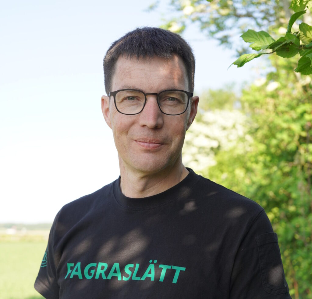 Per Modig, växtodlingsstrateg, Ekologiska Lantbrukarna. Halvfigur i halvskugga av en buske. på en svart t-shirt står det Fagraslätt med gröna bokstäver.