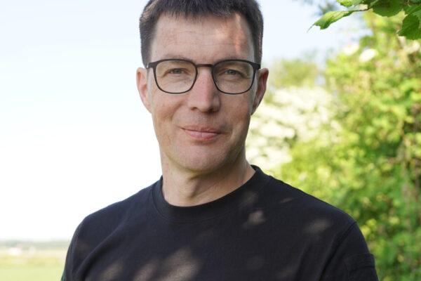 Per Modig, växtodlingsstrateg, Ekologiska Lantbrukarna. Halvfigur i halvskugga av en buske. på en svart t-shirt står det Fagraslätt med gröna bokstäver.